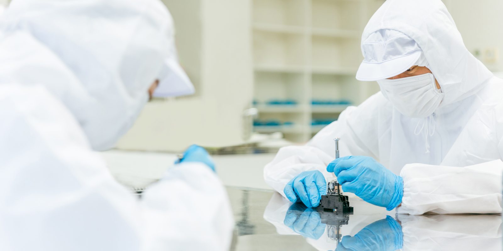 Two technicians in cleanroom coveralls working on a component at a bench.