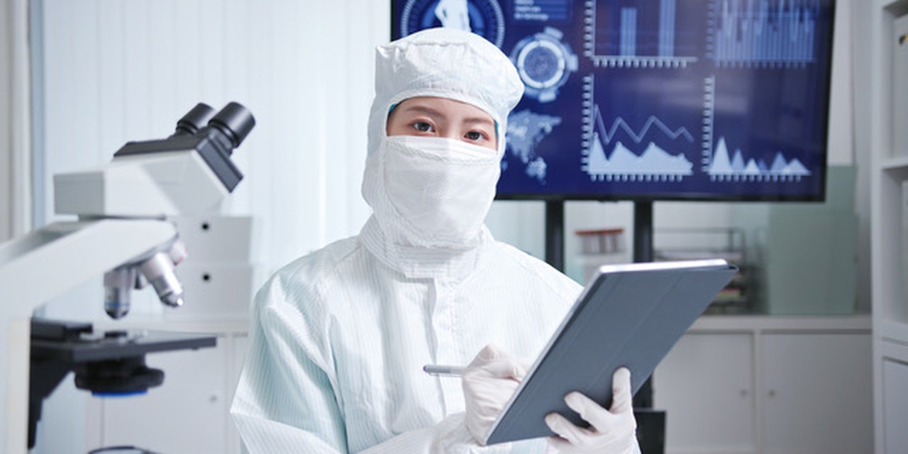 A cleanroom technician with a tablet, a microscope and process monitors behind them.