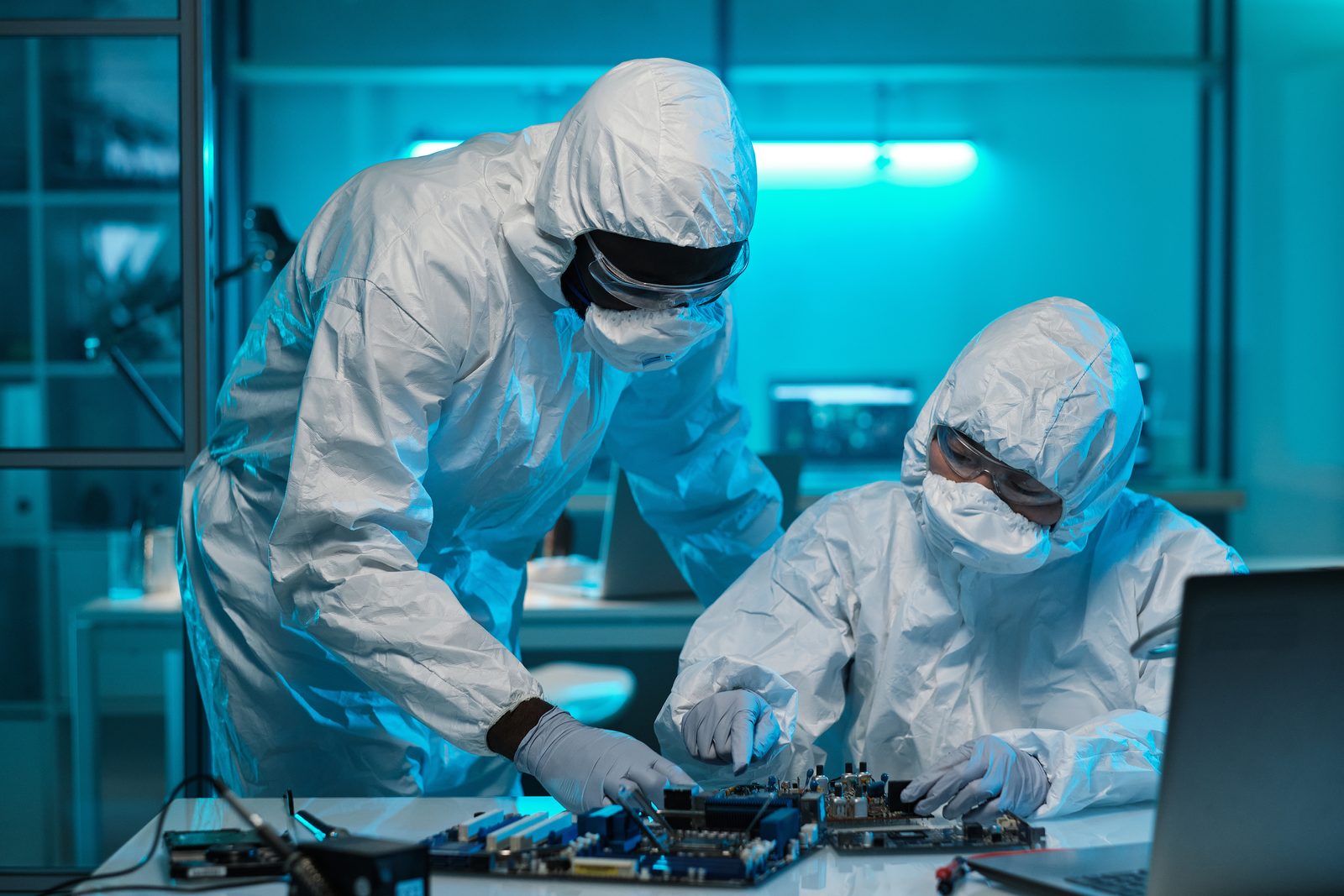 Two technicians in cleanroom suits inspecting a circuit board under blue cleanroom lighting.